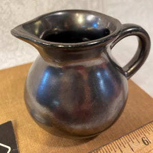 May include: A small, round, black ceramic pitcher with a glossy, metallic finish, a curved handle, and a pouring spout. A wooden ruler is visible at the base, showing the pitcher is approximately 5 inches wide at its widest point.