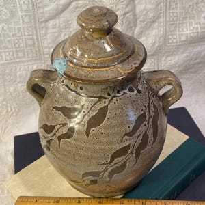 May include: A handcrafted ceramic jar with a lid, featuring a speckled tan and brown glaze. The jar has two handles and is decorated with a leaf pattern. The lid has a small knob. A ruler is at the bottom of the image.
