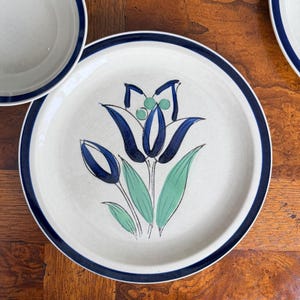 May include: White ceramic plate with a blue floral design and a dark blue rim. The floral design features blue tulip-like flowers and green leaves. The plate is part of a dinnerware set.