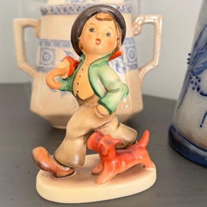 May include: A ceramic figurine of a young boy in a green jacket, brown pants, and a black hat, walking with a red dog. The figurine has a cream-colored base and is set against a blurred background of other ceramic items.