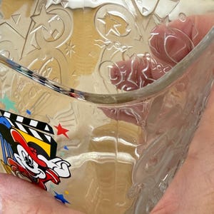 以下が含まれることがあります： 星とミッキーマウスのデザインがエンボス加工された透明なガラスボウル。ボウルには、フィルムストリップと赤い星が付いた、ミッキーマウスのカラフルなグラフィックが側面にあります。ボウルは人が持っています。