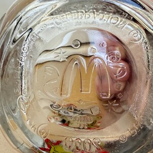 以下が含まれることがあります： 透明なガラス製のマクドナルド記念グラス。グラスの底には、マクドナルドのロゴ、星、そして「Celebration 2000」の文字が描かれています。