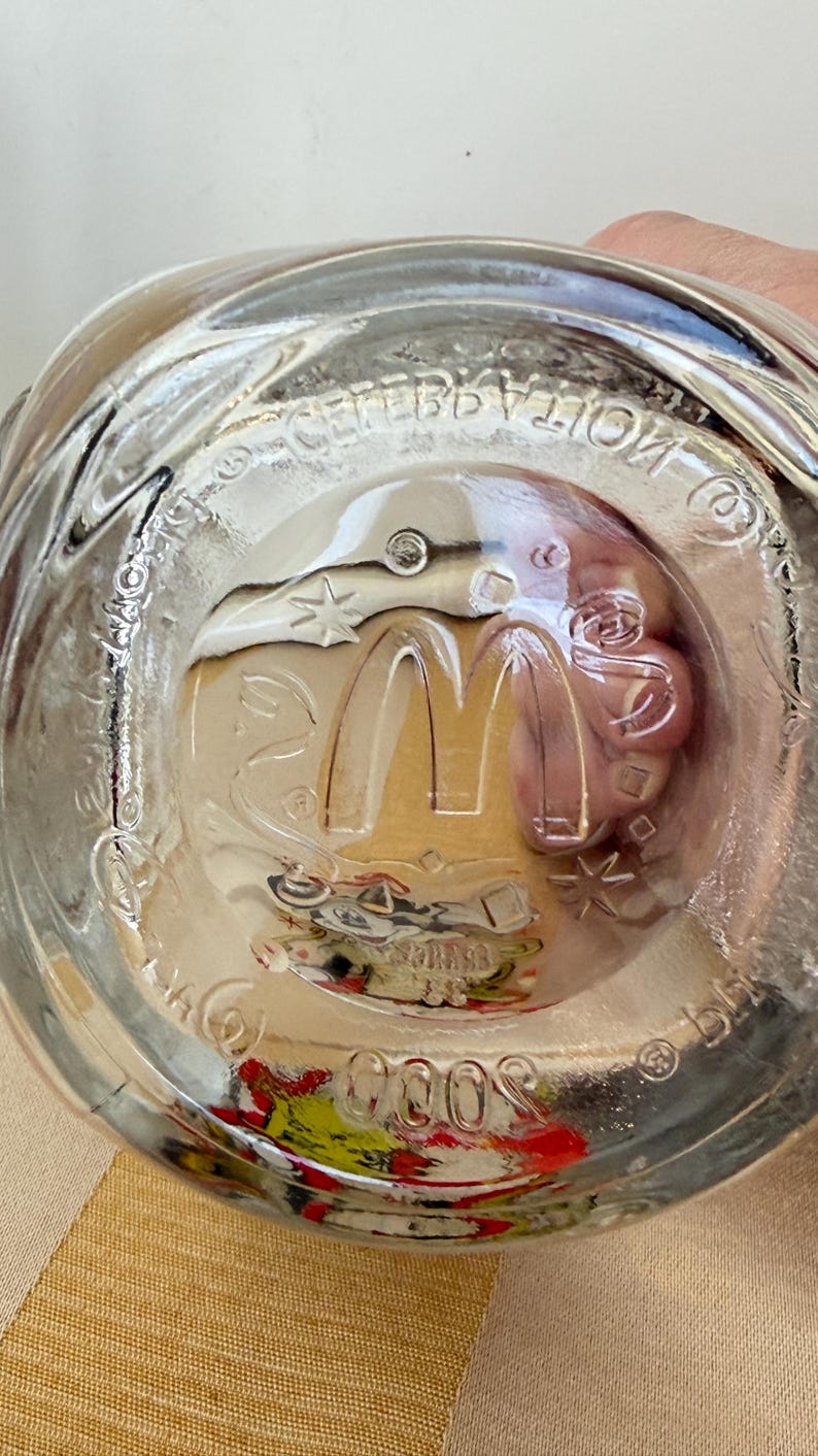 以下が含まれることがあります： 透明なガラス製のマクドナルド記念グラス。グラスの底には、マクドナルドのロゴ、星、そして「Celebration 2000」の文字が描かれています。