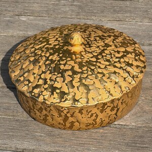 May include: A round ceramic lidded container featuring a metallic gold finish with a textured, mottled pattern across the surface. The lid includes a small, rounded central knob, and the overall design displays a reflective, uneven surface texture.