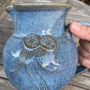 May include: A handmade blue ceramic pitcher with a handle and spout. The pitcher features two circular, embossed designs and vertical lines. The glaze has a speckled appearance with brown accents.