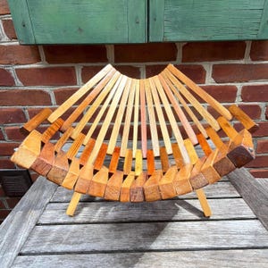 May include: A collapsible wooden fruit basket constructed from multiple light and medium brown slats arranged in a fan-like pattern. The structure features a curved, open design with a central base and two small legs for stability on a flat surface.