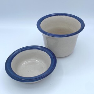May include: Two ceramic bowls with a cream-colored interior and a dark blue rim. One bowl is a small, shallow dish, and the other is a larger, cylindrical pot. The bowls are likely handmade and have a rustic aesthetic.