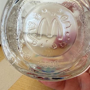 以下が含まれることがあります： 底にエンボス加工されたマクドナルドのロゴが入った透明なガラスのコップ。ロゴは象徴的なゴールデンアーチです。ガラスはわずかにテクスチャ加工された表面で、人が持っています。