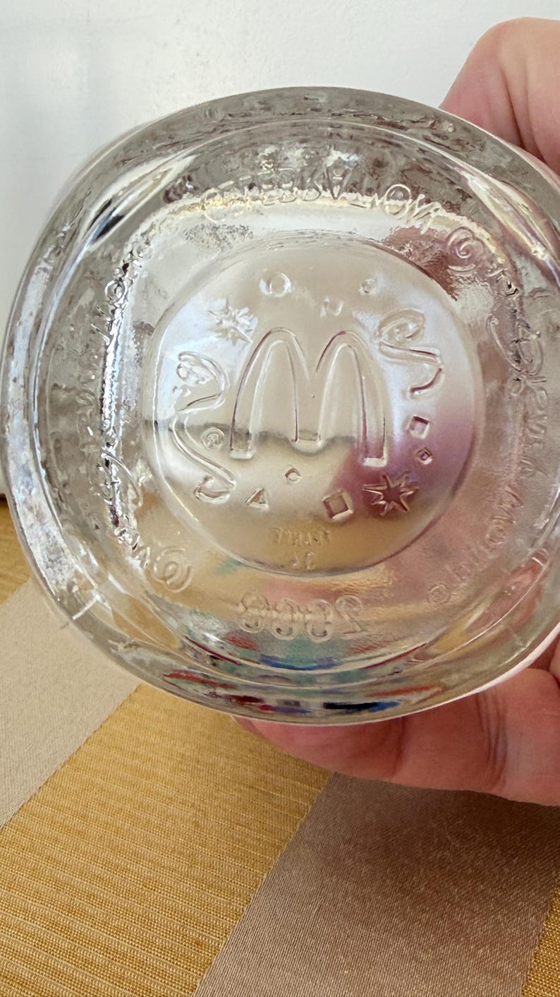 以下が含まれることがあります： 底にエンボス加工されたマクドナルドのロゴが入った透明なガラスのコップ。ロゴは象徴的なゴールデンアーチです。ガラスはわずかにテクスチャ加工された表面で、人が持っています。