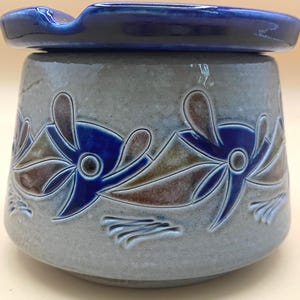 May include: A ceramic jar with a lid, featuring a blue and brown floral design. The jar is a light gray color with a dark blue lid. The floral design is repeated around the jar's body. The jar has a small handle on the lid.