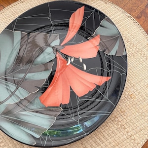 May include: Black ceramic plate with a floral design. The plate features a large coral-colored flower with white accents and gray-green leaves. The background is black with a web-like pattern. The plate is on a woven mat.
