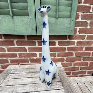 May include: A tall, slender ceramic giraffe figurine with a white base and a repeating pattern of dark blue, hand-painted stars. The giraffe has a long neck, small ears, and two small ossicones on top of its head, featuring a smooth, glossy finish.
