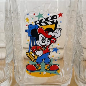 以下が含まれることがあります： 映画をテーマにした、カラフルなミッキーマウスのデザインが施された透明なグラス。ミッキーは赤い帽子とジャケット、青いズボン、赤い靴を履いています。グラスには2000の刻印とディズニーのロゴが入っています。