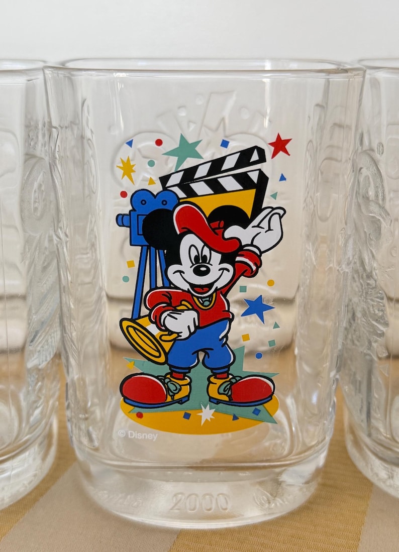 以下が含まれることがあります： 映画をテーマにした、カラフルなミッキーマウスのデザインが施された透明なグラス。ミッキーは赤い帽子とジャケット、青いズボン、赤い靴を履いています。グラスには2000の刻印とディズニーのロゴが入っています。
