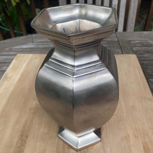 May include: A silver-colored vase with a geometric design. The vase has a hexagonal opening and base, with a rounded body and a series of horizontal lines near the top. The vase is made of metal and sits on a wooden surface.