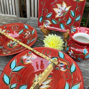 May include: A collection of red ceramic serving dishes with floral designs in white, blue, and yellow. The set includes a square platter, bowls, and a vase. A wooden ruler measures the diameter of a bowl, approximately 12 inches.