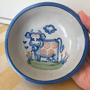May include: A ceramic bowl with a blue rim and a hand-painted cow design. The cow is blue with pink spots and is surrounded by blue hearts, clouds, and flowers. The bowl is white on the inside.