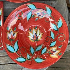 May include: A large, round, red ceramic plate with a floral design. The flowers are white, yellow, and red, with blue leaves and stems. The plate has a spiral pattern and is likely for serving food.