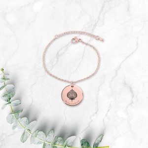 May include: Rose gold bracelet with a circular pendant. The pendant features a stylized shell design and the words "Buen Camino". The bracelet is displayed on a white marble surface with green foliage.