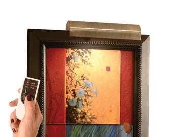May include: A bronze-colored picture light is mounted above a framed painting. The painting features a floral design with blue and white flowers, set against a warm orange and red background. A remote control with the text "1 Concept" is held in the frame.