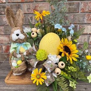 May include: Easter centerpiece featuring a straw bunny holding a basket of eggs, a large yellow egg, sunflowers, and decorative ribbon with bunny prints. The arrangement is set on a wooden board.