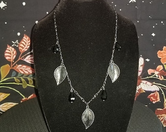May include: A silver necklace with black leaf charms and black teardrop beads. The necklace is displayed on a black velvet mannequin bust against a black background with pink moons and white stars.