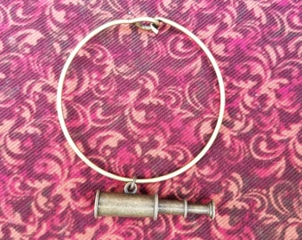 May include: A gold-toned bangle bracelet with a small, antique-looking telescope charm hanging from it. The bracelet is lying on a pink and brown patterned fabric.