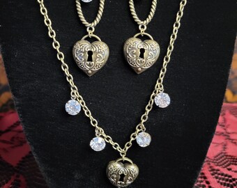 May include: A gold-tone necklace and earrings set featuring a heart-shaped pendant with a keyhole design and clear crystal accents. The necklace has a delicate chain with dangling crystals.
