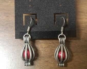 May include: A pair of silver-colored earrings with a cage-like design, each holding a red bead. The earrings are displayed on a black card that reads "FASHION EARRINGS" in gold lettering.