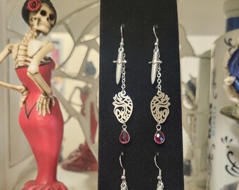 May include: A pair of silver earrings with a heart-shaped charm, a dagger charm, and a red gemstone dangle. The earrings are displayed on a black background.