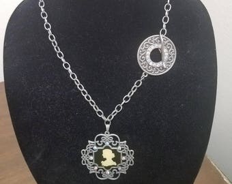 May include: A silver chain necklace with a cameo pendant featuring a woman's silhouette. The pendant is surrounded by a decorative frame with small rhinestones. The necklace also has a circular charm with rhinestones.