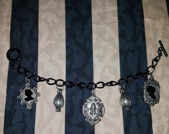 May include: A black chain bracelet with five silver charms. The charms include a fleur-de-lis, two cage-like charms with a pearl inside, and two silhouette charms of a person's head.
