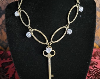 May include: A gold-toned necklace with a key pendant and clear crystal accents. The necklace features a chain with oval links and dangling crystals.
