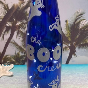 Op de afbeelding: Een blauwe glazen fles met de woorden "the BOO crew" in zilveren letters. De fles is versierd met zilveren spookfiguren en kleine witte lichtjes. De fles heeft een wit en zilver geweven bovenkant en staat tegen een tropische achtergrond.