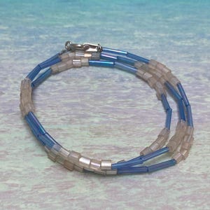Op de afbeelding: Een kralenarmband met een zilveren sluiting. De armband heeft drie strengen kleine, lichtblauwe en heldere kralen. De kralen zijn aan elkaar geregen om een flexibel, cirkelvormig ontwerp te vormen. De armband wordt getoond op een lichtblauwe en witte ondergrond.