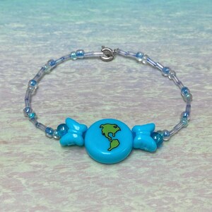 May include: A beaded bracelet featuring a light blue charm with a green map of the world. The bracelet has clear and blue beads with a silver clasp. Two light blue butterfly-shaped beads flank the central charm.