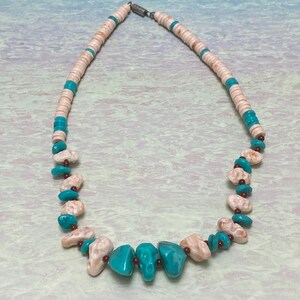 Op de afbeelding: Een turquoise en beige kralenketting. De ketting heeft een verscheidenheid aan kralen, waaronder turkooizen stenen, beige schelpachtige kralen en kleine rode kralen. De ketting is geregen aan een zilverkleurige ketting.