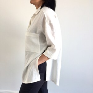 May include: A white linen shirt with a long sleeve and a single patch pocket on the left side. The shirt has a high-low hemline and is worn with black pants.