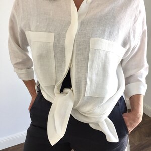 May include: A white linen shirt with long sleeves and two large pockets. The shirt is tied at the waist.