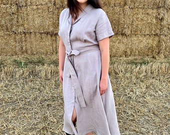 May include: A light grey linen dress with short sleeves, a collared neckline, and a tie belt. The dress is knee-length and buttoned down the front. The person is wearing black sandals. The background is a stack of hay bales.