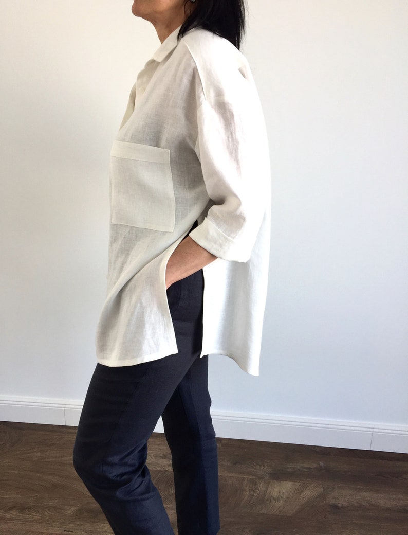 May include: A white linen shirt with a long sleeve and a single patch pocket on the left side. The shirt has a high-low hemline and is worn with black pants.
