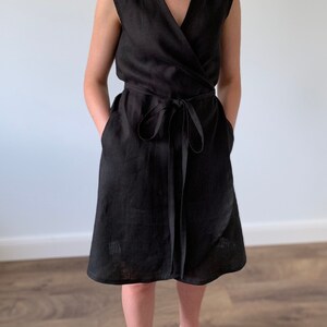 May include: A black linen wrap dress with a V-neckline and short sleeves. The dress has a tie belt at the waist and pockets on the sides.