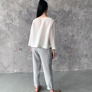 May include: A white linen top with long sleeves and a relaxed fit, paired with light gray linen pants. The model is wearing gold sandals.