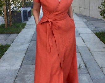 May include: A woman wearing a long, rust-colored wrap dress with a self-tie belt. She is standing on a paved walkway with green plants in the background.