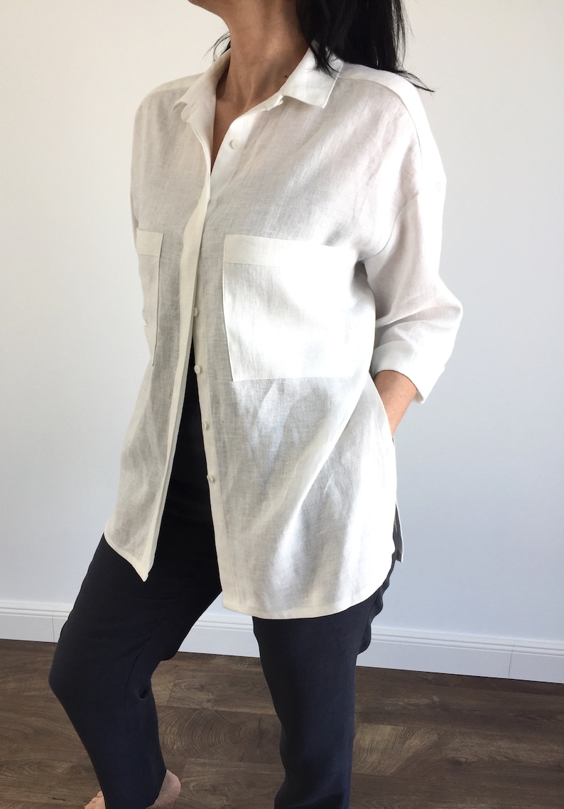 May include: A white linen shirt with a long sleeve and a single pocket on the left side. The shirt is unbuttoned and the model is wearing black pants.