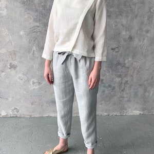 May include: A white linen top with a diagonal seam and a tie waist, paired with light gray linen pants and gold sandals.