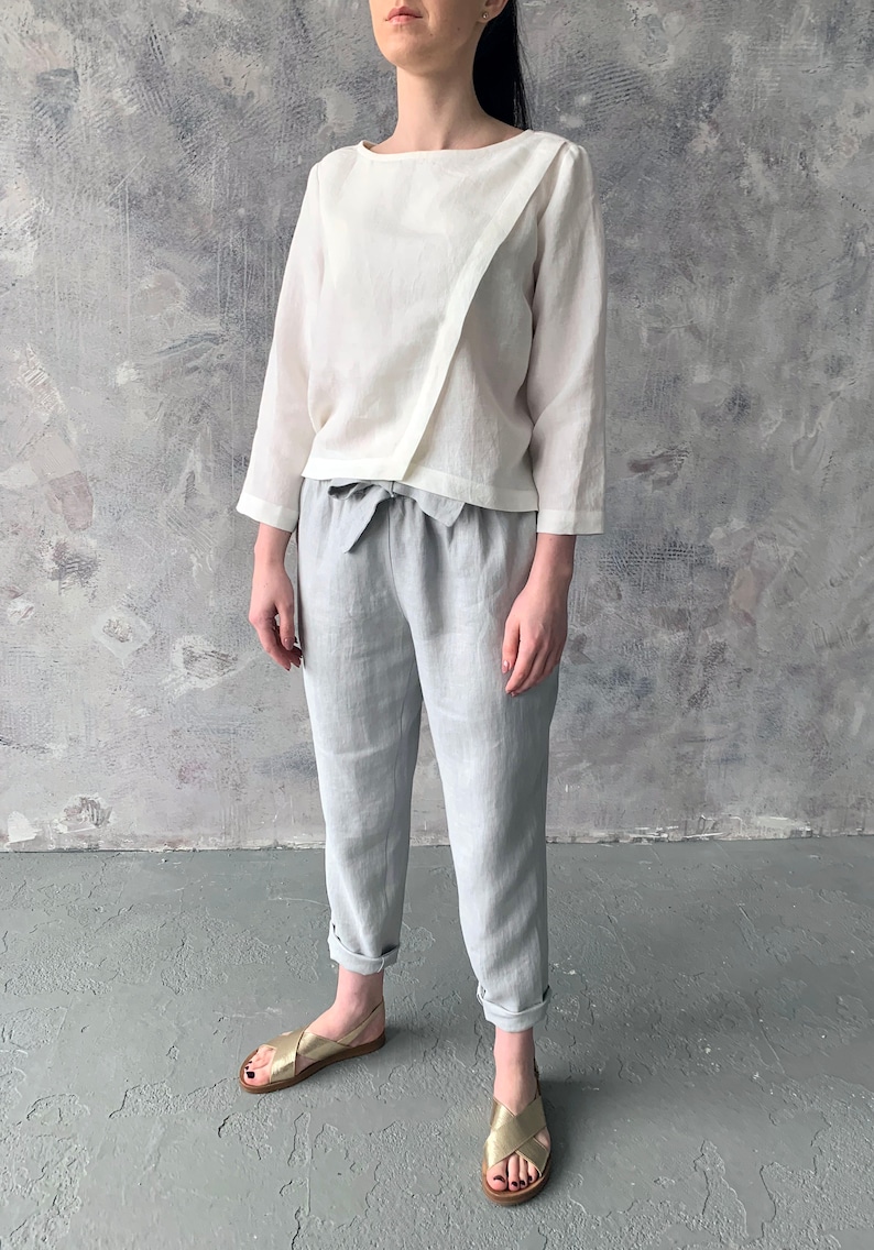 May include: A white linen top with a diagonal seam and a tie waist, paired with light gray linen pants and gold sandals.