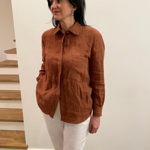 Op de afbeelding: Een roestkleurig linnen overhemd met een knoopsluiting en lange mouwen. Het overhemd heeft een borstzak en wordt gedragen met een witte linnen broek.