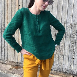 May include: A woman wearing a green long-sleeved linen shirt with a button placket and mustard yellow linen pants.