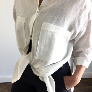May include: A white linen shirt with long sleeves and two large pockets. The shirt is tied at the waist and worn with black pants.